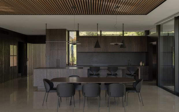 A dark, refined open-plan dining and kitchen interior. A long timber dining table with six upholstered chairs sits in the foreground, while a minimalist kitchen with a large island, integrated dark cabinetry, stone surfaces and bar seating occupies the background. Floor-to-ceiling glazing brings in filtered greenery, and a slatted timber ceiling adds warmth and texture. Pendant lights hang above both the island and dining area.