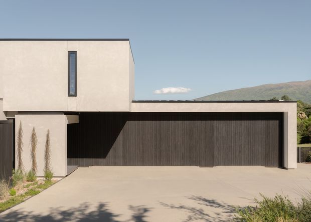 Front exterior view of a contemporary flat-roofed residence with a minimalist pale rendered upper volume, a deep recessed entry or carport zone, and a wide dark timber-look garage door. A broad concrete driveway dominates the foreground, with sparse landscaping and a mountainous backdrop.
