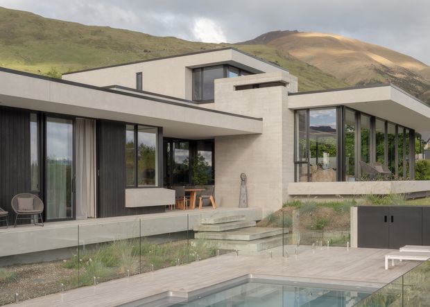 Exterior view of a contemporary multi-level hillside residence with strong horizontal flat-roofed forms, extensive floor-to-ceiling glazing, dark vertical cladding, pale masonry walls, and a sheltered outdoor entertaining area. A glass pool fence encloses a timber deck and swimming pool in the foreground, with rugged green and ochre mountain terrain behind the house.