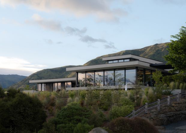 A low-slung contemporary residence set into a rugged, landscaped mountain site. The home features broad floor-to-ceiling glazing, dark horizontal window frames, layered flat roof planes, and an understated palette of grey and charcoal materials. Native-style planting, stone retaining walls, and timber fencing surround the building, with steep mountainous terrain visible behind it.