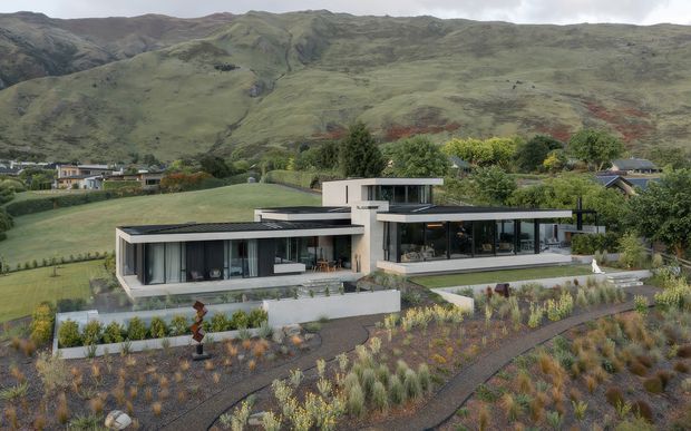 A contemporary single-storey residence set within a landscaped hillside site, viewed from above. The home has long, low horizontal forms, extensive floor-to-ceiling glazing, flat roofs, pale rendered walls, dark window frames, and generous terraces. A planted garden and curved pathways occupy the foreground, while dramatic green mountains rise behind the property.