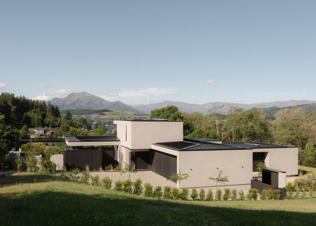 A low, modern residential house set into a landscaped sloping site, viewed from across a lawn. The home has minimalist rectangular volumes, flat roof forms with dark metal roofing, smooth pale rendered walls, large dark-framed glazed openings, and landscaped garden beds. A broad mountain and forested landscape forms the background.