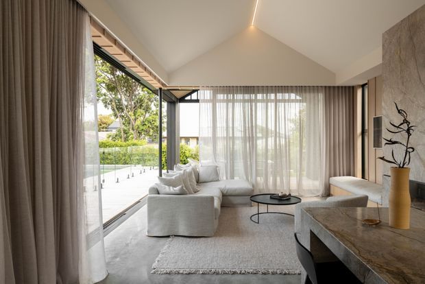 Bright contemporary living room with a high pitched ceiling, floor-to-ceiling glazing, sheer curtains, and an open corner connection to an outdoor terrace. The room features a pale sectional sofa, textured area rug, round black coffee table, sculptural side table, and a substantial stone-topped built-in console or counter along the right wall. Neutral finishes and concealed linear lighting create a calm, refined atmosphere.