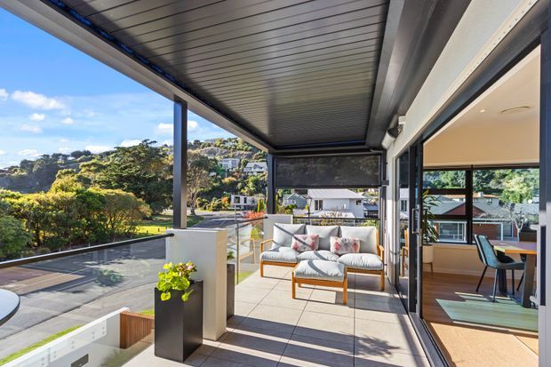 A bright contemporary covered terrace overlooking a landscaped suburban setting, with slim dark structural posts, a slatted metal soffit, glass balustrading, pale tiled paving, outdoor lounge furniture, and large sliding glass doors opening into an adjacent dining area. The terrace connects directly to the interior and benefits from expansive views and abundant natural light.