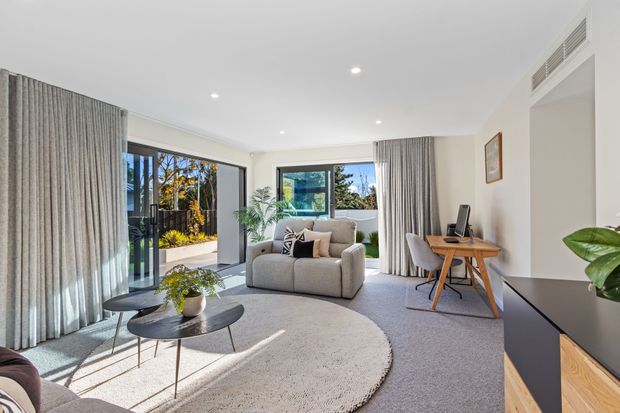 Bright contemporary living room with large sliding glass doors opening to a landscaped outdoor area. The room features a light neutral palette, pale carpet, floor-to-ceiling grey curtains, a rounded cream sofa and armchair, nesting coffee tables, a compact timber desk, and a dark built-in media cabinet. Recessed ceiling lights and concealed air-conditioning vents contribute to a clean, refined interior.