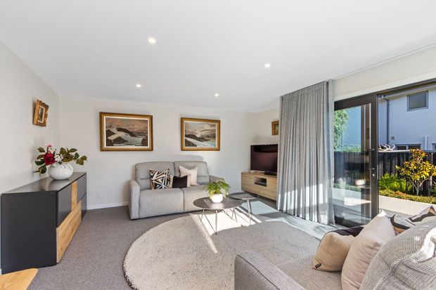 Bright contemporary living room with a grey sectional sofa, round textured rug, timber-and-black media cabinetry, framed artwork, and full-height sliding glass doors opening to an outdoor area. Sheer curtains and recessed ceiling lights create an airy, polished atmosphere.