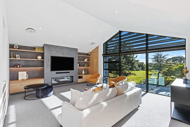 Bright contemporary living room with a large white sectional sofa, round coffee tables, built-in timber shelving, and a dark stone or concrete fireplace and TV feature wall. Floor-to-ceiling black-framed glazing opens to a leafy garden view, while warm timber wall panelling and recessed lighting add contrast to the light neutral interior.
