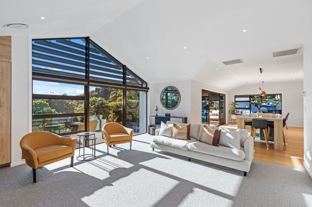 Bright contemporary open-plan living room with a large light-grey sectional sofa, two ochre lounge chairs, and a small round side table. Floor-to-ceiling glazing with horizontal external louvres brings in strong sunlight and views of trees. The space connects to a dining area and kitchen with timber flooring, white walls and ceiling, recessed downlights, and a round feature window.