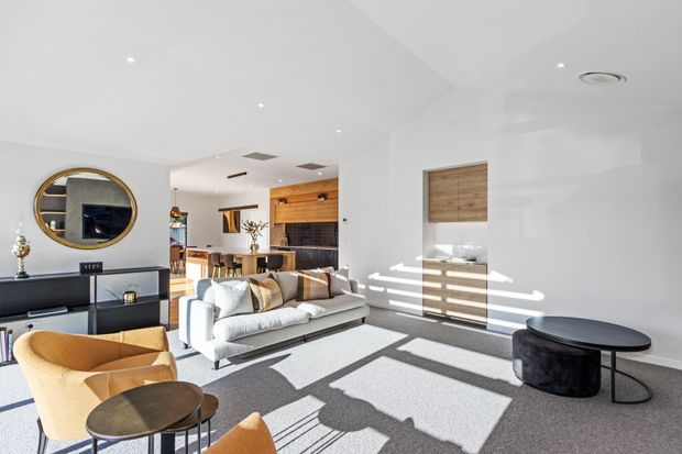 Bright open-plan living room with a contemporary, minimalist fit-out. The space features a large light-coloured sofa, sculptural mustard lounge chairs, round coffee tables, a black low media console with circular mirror above, pale carpet, white walls and ceiling, and timber kitchen cabinetry visible in the background. Strong sunlight creates geometric shadows across the floor and walls.