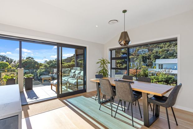 Bright contemporary dining area opening onto a balcony or terrace through large sliding glass doors. The room features a timber dining table with upholstered dining chairs, light timber flooring, white walls and ceiling, dark-framed glazing, a pendant light, and leafy residential views beyond.