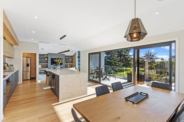Bright open-plan kitchen and dining area with warm timber flooring and cabinetry, a large marble-look island, dark built-in shelving, and extensive floor-to-ceiling glazing opening to an outdoor terrace and garden. A timber dining table sits beneath a geometric pendant light, creating a contemporary, polished family entertaining space.