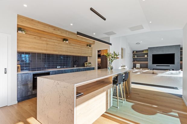 Bright open-plan kitchen and living area with a large waterfall-edge stone island, warm timber cabinetry and flooring, dark blue tiled splashback, and integrated black linear pendant lighting. A living room with built-in shelving, television, and light sofa is visible beyond the kitchen.