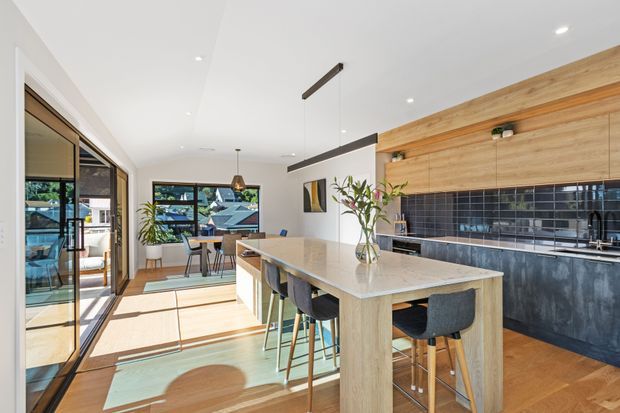 Bright contemporary kitchen and dining area with a large light-stone island, timber cabinetry and wall panelling, dark tiled splashback, integrated appliances, and bar seating. Full-height glazing and sliding doors open the space toward an outdoor view, while pendant and recessed lighting create an airy, refined interior.
