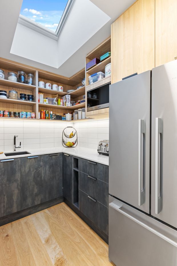 Compact, highly functional kitchen pantry with L-shaped dark cabinetry, pale engineered-looking benchtops, white tiled splashback, open timber shelving filled with pantry items, integrated appliances, and a large stainless-steel refrigerator. A roof skylight brings bright natural light into the space.