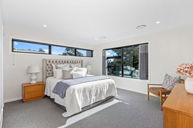 Bright contemporary bedroom with a large upholstered button-tufted bedhead, layered white bedding, grey carpet, timber bedside table and dresser, and expansive corner glazing overlooking greenery. The room has recessed ceiling lights, simple white walls, and a calm, refined neutral palette.