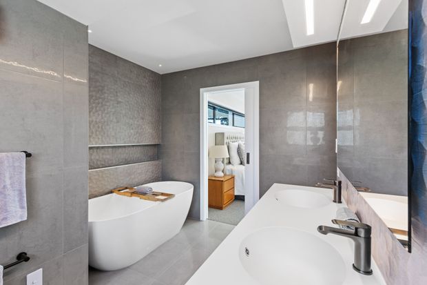 Contemporary bathroom with a freestanding white bathtub, double vanity basins, large grey tiled walls and floor, a mirrored wall, and bright linear ceiling lighting. A bedroom is visible through the open doorway.