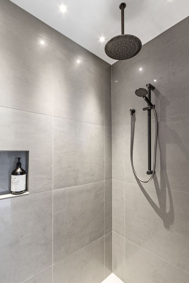 A minimalist walk-in shower with large-format light grey wall tiles, a dark metal ceiling-mounted rainfall showerhead, matching handheld shower, recessed niche, and recessed ceiling spotlights. The compact space has a clean, contemporary finish with strong geometric tile lines and high contrast from the dark fixtures.