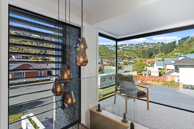 Bright contemporary interior overlooking a suburban hillside neighbourhood through expansive corner glazing. The space features a stair or landing with a clear glass balustrade, dark-framed windows, a textured dark stone feature wall, a timber ledge, and sculptural pendant lights. Outdoor views and neighbouring houses are visible beyond the glazing.