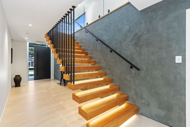 Bright contemporary hallway featuring a floating timber staircase with warm under-step lighting, slim black vertical balustrade rods, and a black handrail mounted against a textured grey feature wall. Pale flooring and white walls create a clean, modern entry sequence, with glazing visible at the upper landing.