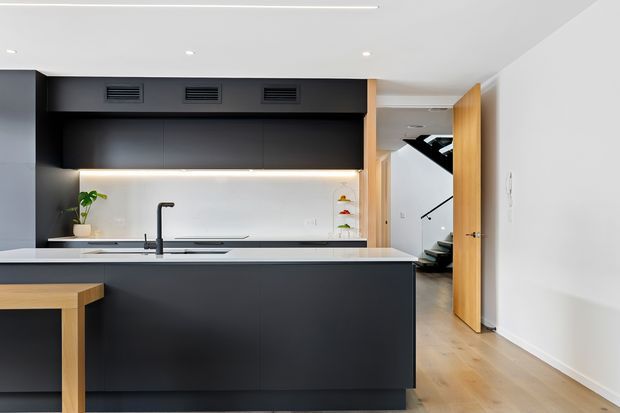 Contemporary minimalist kitchen with a large dark island and black cabinetry, pale benchtops, integrated appliances, and a warm timber stool or breakfast ledge. A timber door opens to a hallway and black staircase beyond. The room has a clean white ceiling with recessed lighting and light timber flooring.