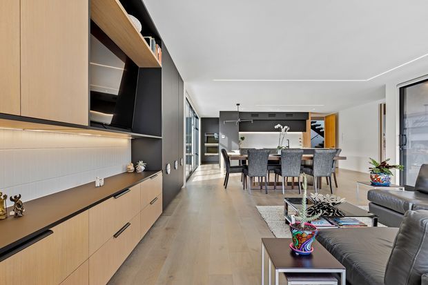 Open-plan contemporary interior featuring a built-in timber-toned media and storage wall, dark cabinetry, a dining area, and an adjoining living room. The space has pale timber flooring, white walls and ceiling, integrated linear lighting, large glazing, and a neutral palette with colourful decorative accents.