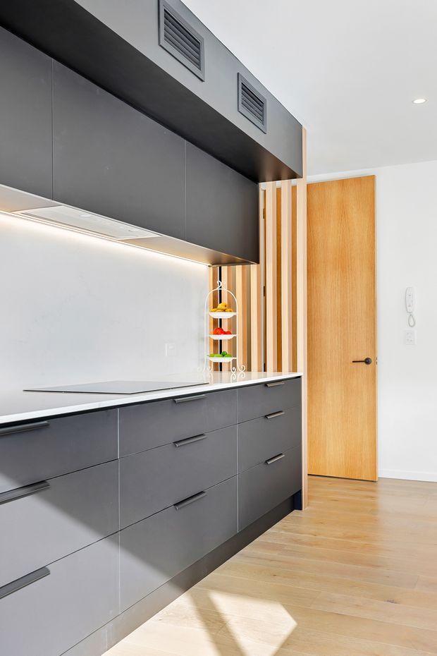A compact, contemporary kitchen with matte dark-grey flat-panel cabinetry, integrated upper storage and under-cabinet lighting. The kitchen has a pale stone or composite benchtop, a light neutral splashback, and a light timber floor. A vertical timber slat screen and matching timber door add warmth, while exposed ventilation grilles and recessed ceiling lighting contribute to the clean modern fit-out.