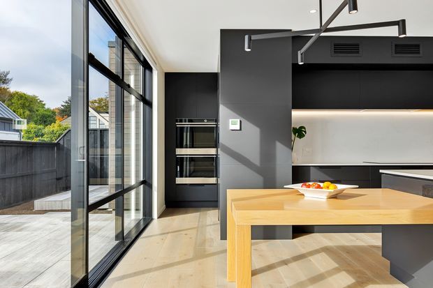 Bright contemporary kitchen with a large timber island and integrated seating, matte black cabinetry, built-in appliances, a pale stone or concrete-look backsplash, and expansive black-framed glazing opening to an outdoor deck. Linear pendant lighting and concealed under-cabinet illumination create a clean modern fit-out.