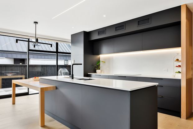 Contemporary open-plan kitchen with a large matte dark-grey island, white stone benchtop, integrated black cabinetry, illuminated white splashback, timber breakfast bar, and full-height black-framed glazing. Vertical timber screening adds warmth at the right edge, while a dining area and covered exterior space are visible beyond the kitchen.
