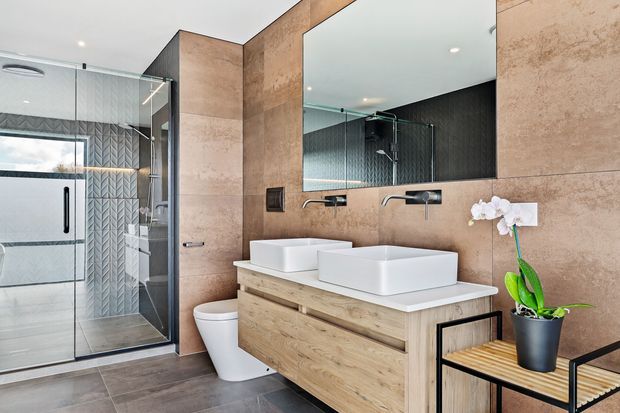 Contemporary bathroom featuring a double vanity with wall-mounted black tapware, timber-look drawer cabinetry, large-format beige stone-look wall tiles, a frameless glass shower enclosure, patterned shower feature tiles, and a freestanding toilet. A large mirror and orchid add visual interest.