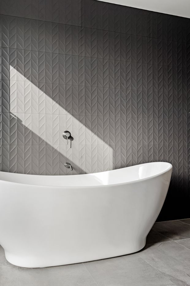 A contemporary bathroom featuring a freestanding white sculptural bathtub against a dark grey geometric textured feature wall. Minimal chrome wall-mounted plumbing fixtures and a strong diagonal beam of natural light create contrast, while large grey floor tiles complete the monochromatic scheme.