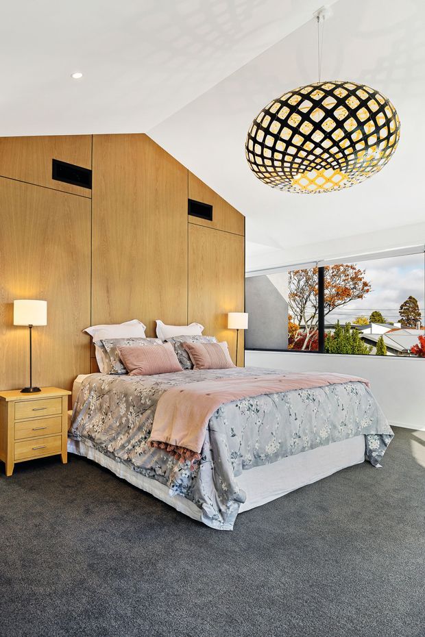 A contemporary bedroom with a large bed dressed in soft blue-grey and blush floral bedding, a full-height warm timber feature wall with integrated dark vents, matching timber bedside storage, and a sculptural black-and-gold pendant light. A broad window overlooks suburban rooftops and trees, while grey carpet covers the floor.