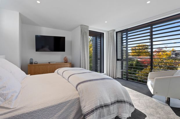 Bright contemporary bedroom with a large bed, white bedding, grey carpet, a timber media console and wall-mounted television. Floor-to-ceiling glazing with dark horizontal louvres provides extensive views to trees outside, while sheer curtains soften the windows. A white sculptural lounge chair sits beside the glazing.