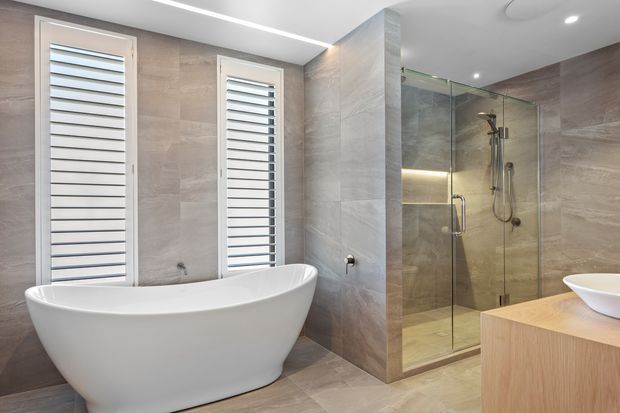 Contemporary bathroom featuring a freestanding white bathtub beneath two narrow shuttered windows, a frameless glass walk-in shower with wall-mounted fixtures, large-format beige-grey stone-look tiles, recessed ceiling lighting, and a light timber vanity with a white vessel basin.