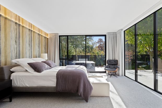 Bright, contemporary bedroom with a large upholstered bed, neutral bedding, and a soft grey carpet. A dramatic vertical timber feature wall runs behind the bed, while floor-to-ceiling black-framed glazing opens the room visually to a leafy outdoor setting. Sheer curtains, a compact modern desk chair, and restrained furnishings create an airy, refined atmosphere.