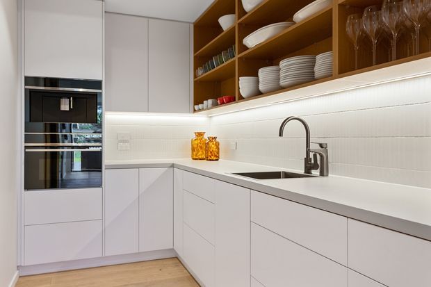Contemporary white kitchen with integrated black appliances, handleless cabinetry, a white benchtop, a stainless steel undermount sink and curved tap, and a textured white tiled splashback. Open walnut-toned shelving displays plates, bowls, glassware and cups, with warm under-shelf lighting. Light timber flooring adds warmth.