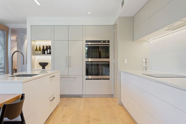 Bright contemporary kitchen with extensive white built-in cabinetry, a central island with sink and gooseneck tap, integrated double ovens, and a long white benchtop along the right wall. The kitchen features pale timber flooring, white tiled splashback, understated recessed lighting, and a partially visible dining area with timber-framed glazing.