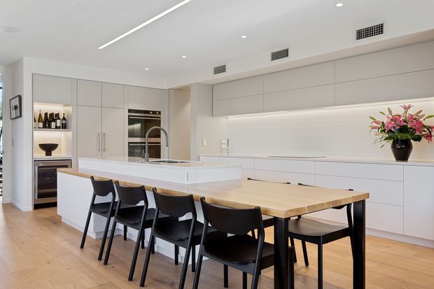 Bright, spacious contemporary kitchen and dining area with extensive matte white cabinetry, integrated appliances, a large white island with a stainless-steel gooseneck tap, and a light timber dining table surrounded by black dining chairs. Light timber flooring, recessed lighting, linear ceiling lighting, and a restrained neutral palette create a clean, refined interior.