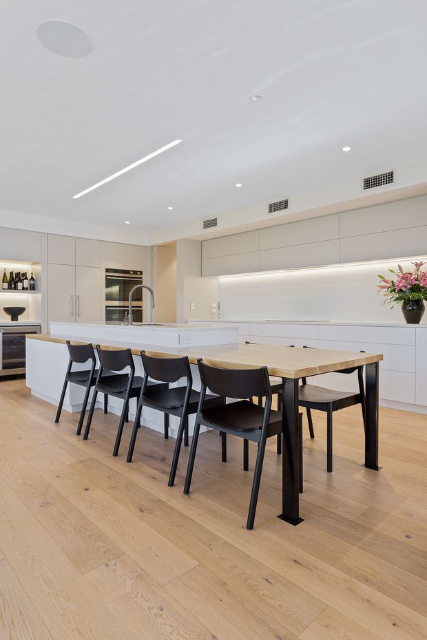 Bright contemporary open-plan kitchen and dining area with streamlined white full-height cabinetry, integrated appliances, a large white island, and a light timber dining table surrounded by black dining chairs. Light oak flooring, concealed under-cabinet lighting, recessed ceiling lights, and linear ceiling lighting create a clean, refined interior.