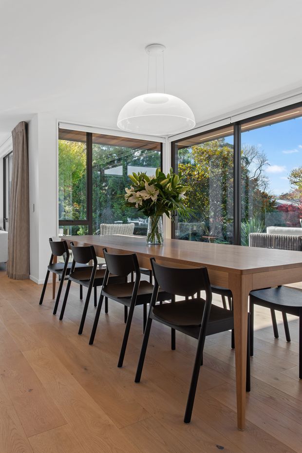 Bright contemporary dining room with a long mid-tone timber dining table and black dining chairs, set against extensive black-framed corner glazing overlooking a leafy garden. The room has light timber flooring, white walls and ceiling, sheer neutral curtains, and a large white pendant light. A floral arrangement forms the table centrepiece.