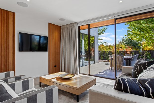 Bright contemporary living room with large sliding glass doors opening to a paved outdoor terrace. The room features a wall-mounted television, vertical timber wall panelling, neutral curtains, a timber coffee table, upholstered sofas and armchairs, and a dining setting visible outside. The space has strong indoor-outdoor connection and abundant natural light.