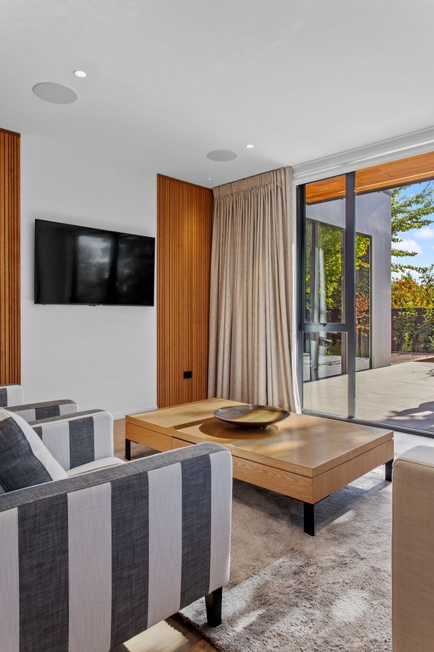 Contemporary living room with grey-and-white upholstered armchairs, a large timber coffee table, wall-mounted television, vertical timber feature panelling, full-height curtains, and expansive glazed doors opening to an outdoor terrace. The room is bright and refined, with a neutral palette and strong indoor-outdoor connection.