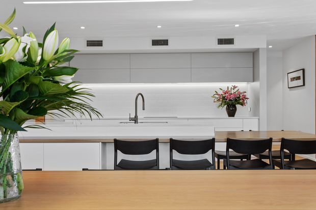 Bright, clean open-plan kitchen and dining area with white flat-panel cabinetry, a long island or counter, integrated appliances, a stainless-steel tap, timber benchtops or dining surfaces, and black dining chairs. Large floral arrangements add a natural accent.