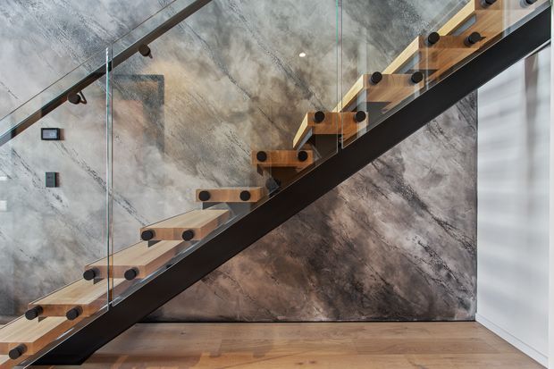 Close-up architectural photograph of a contemporary stairway with floating timber treads mounted on a dark structural stringer, clear glass balustrades, and black circular fixings. The stair is set against a dramatic grey and brown marbled feature wall, with warm timber flooring and a bright window or glazed opening at right.
