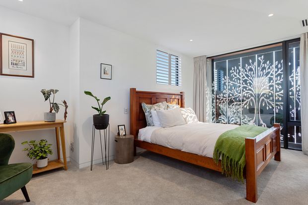 Bright contemporary bedroom with a timber bed, white bedding, grey carpet, indoor plants, simple framed artwork, and a large glazed opening featuring a decorative botanical screen. A slim timber console and armchair complete the calm, natural interior.
