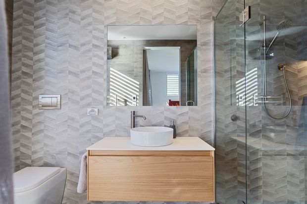 Contemporary bathroom with grey geometric-patterned wall and floor tiles, a wall-mounted timber vanity with a white vessel basin, frameless glass shower enclosure, wall-hung toilet, and large mirror. Natural light enters through louvred windows reflected in the mirror and shower glass.