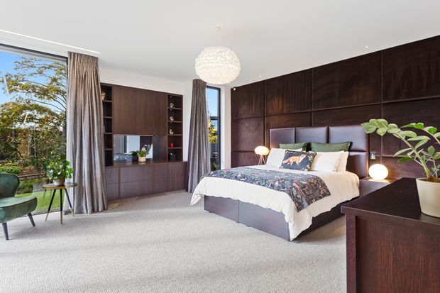 A refined contemporary bedroom with a large upholstered bed set against a dramatic dark timber-panelled feature wall. Matching dark timber built-in cabinetry surrounds a television and shelving, while floor-to-ceiling curtains frame generous glazing. A textured spherical pendant, bedside lighting, indoor plants, and soft neutral carpet create a warm, sophisticated retreat.