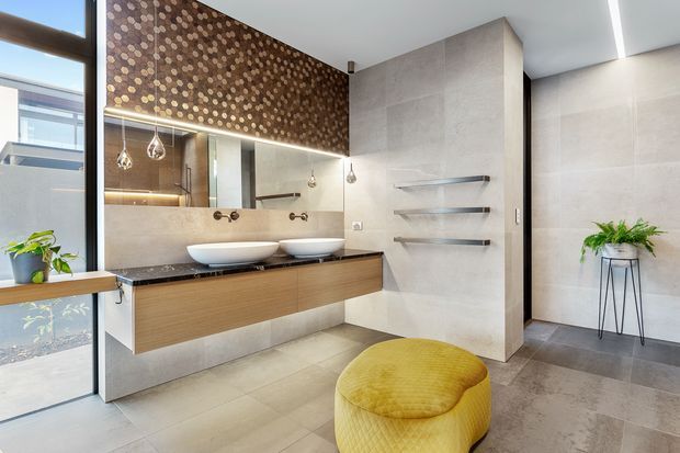 Contemporary bathroom featuring a floating timber vanity with twin white vessel basins, a large mirror, wall-mounted chrome tapware, open shelving, pale stone-look wall and floor tiles, and a decorative dark patterned feature wall above the vanity. A mustard upholstered ottoman and indoor plants add warmth and contrast. Full-height glazing and a linear ceiling light create a bright, refined atmosphere.