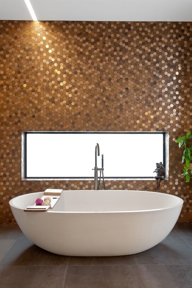 A luxurious contemporary bathroom featuring a freestanding white oval bathtub, wall-mounted chrome tapware, a large horizontal window, and a striking full-height honeycomb mosaic tile wall in warm brown and bronze tones. Large dark stone-look floor tiles and a linear ceiling light complete the refined, spa-like setting.