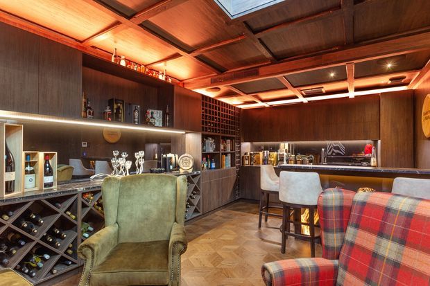 A richly detailed home bar and wine room with dark timber wall panelling and cabinetry, extensive wine storage, a long bar counter with stools, lounge seating, and warm integrated lighting. The ceiling features exposed timber framing and illuminated recessed panels, creating a sophisticated, intimate atmosphere.