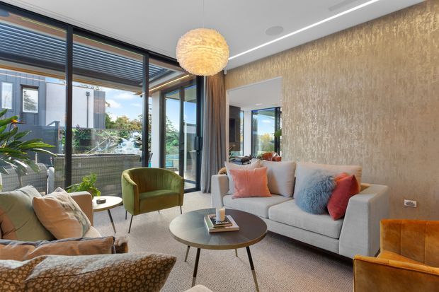 Contemporary living room with floor-to-ceiling black-framed glazing opening to a balcony, a textured feature wall, neutral sectional sofa, velvet accent chairs, patterned armchair, round coffee table, and a sculptural pendant light. The room has a warm, refined palette with layered textiles and abundant natural light.